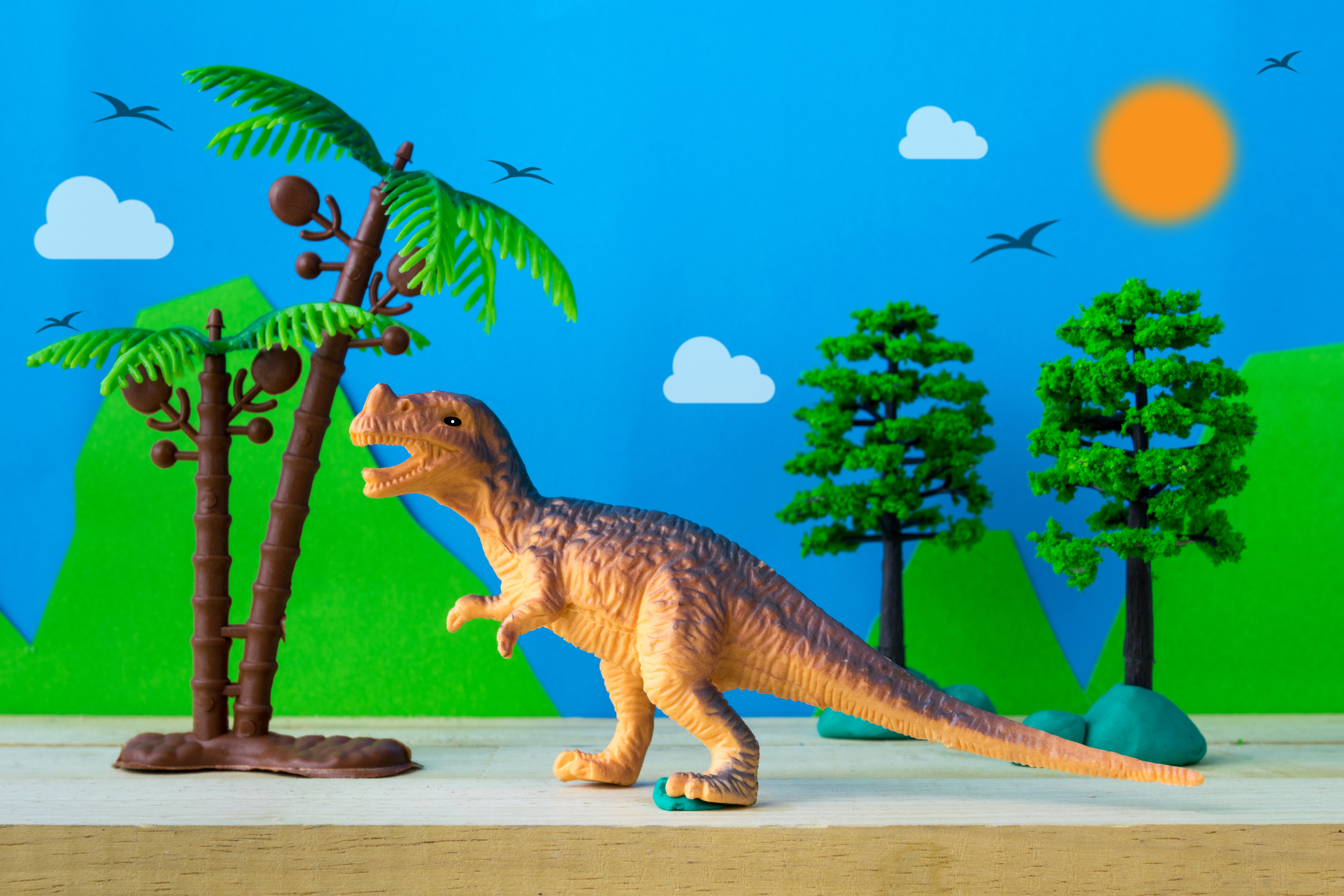 Cover Image: A whimsical diorama featuring a toy T-Rex and toy trees with a paper cutout background featuring mountains, a sky, birds, and a sun.
